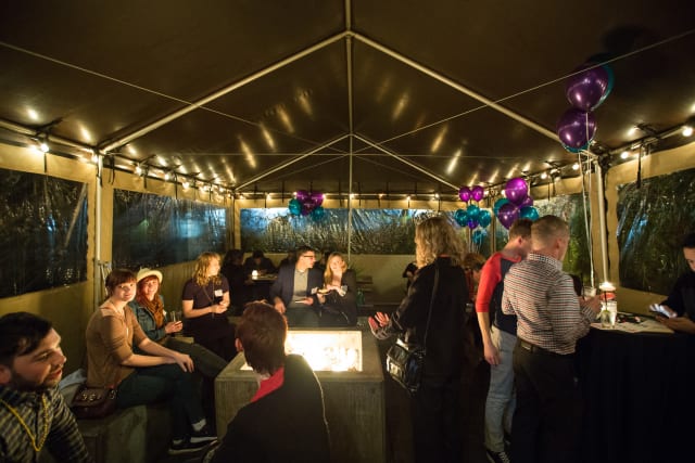 Guests socializing in a tented area with purple balloons, soft lighting, and a central fire pit during the Love Bomb 2017 event supporting My Voice Music.